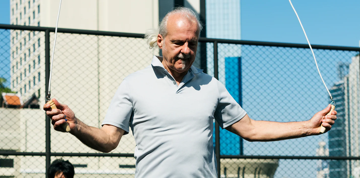 Older man skipping rope outdoors next to a chain-link fence